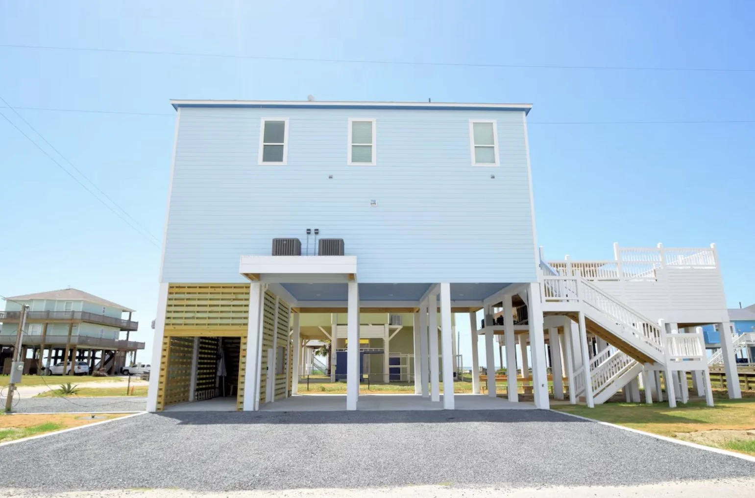 A raised beach house painted light blue with white trim, featuring a staircase on the right side leading to the main entrance and a covered parking area underneath.