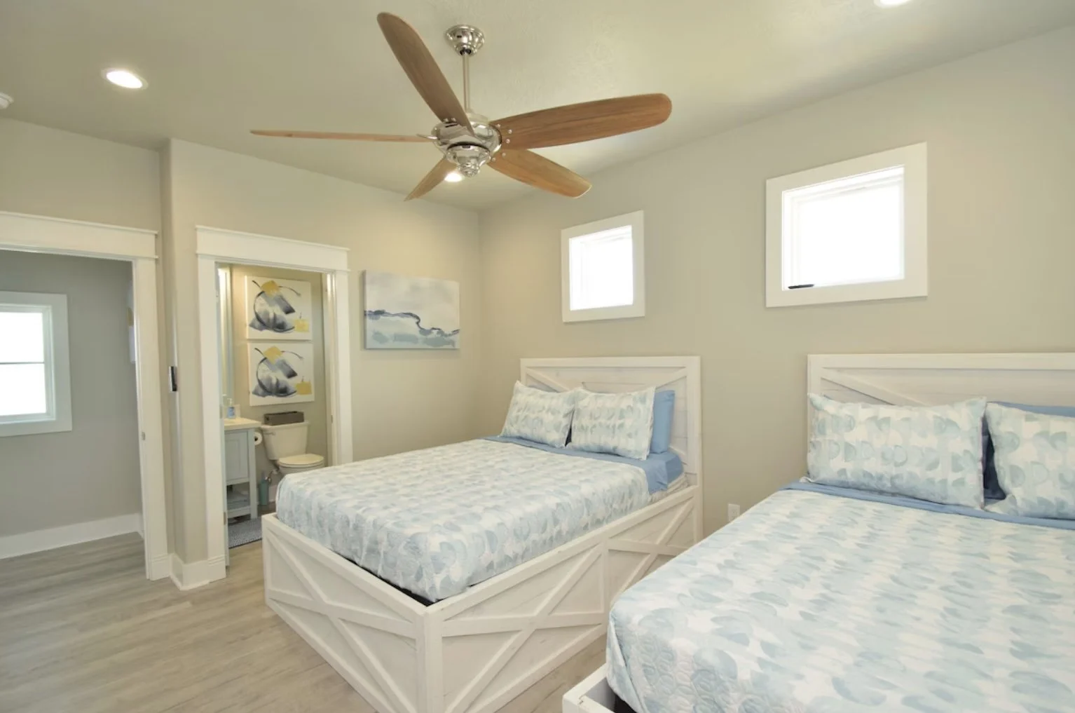 Bedroom with two white wooden beds, a ceiling fan, two small windows, and bathroom with toilet and sink in background.