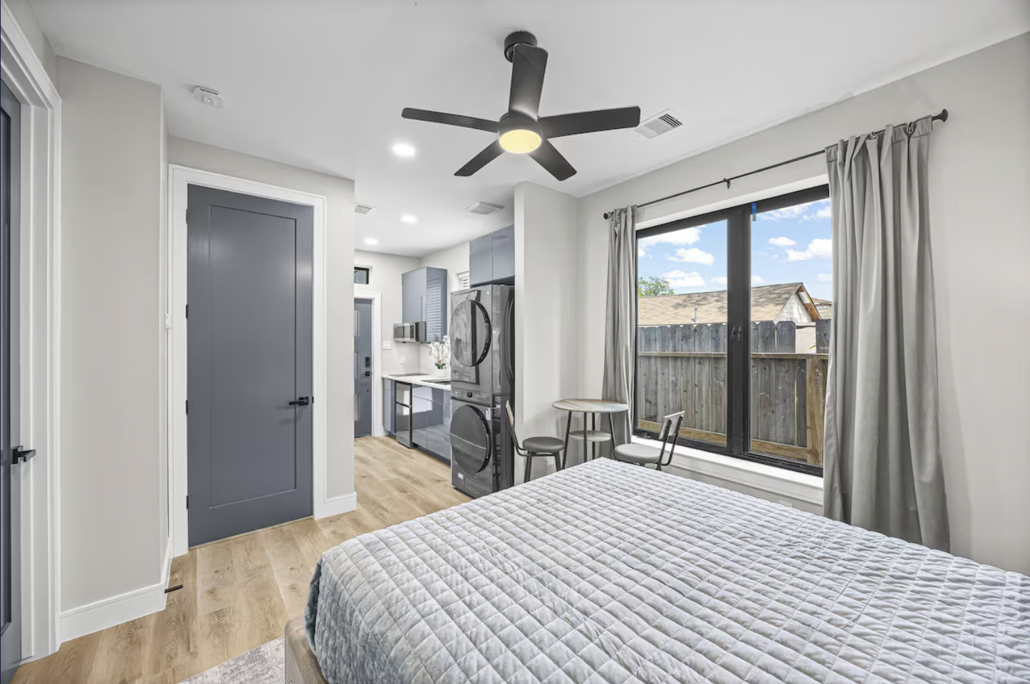 Bright bedroom with a large window, queen bed with light gray quilt, ceiling fan, small round table, chairs, laundry appliances, and a doorway to the hallway.
