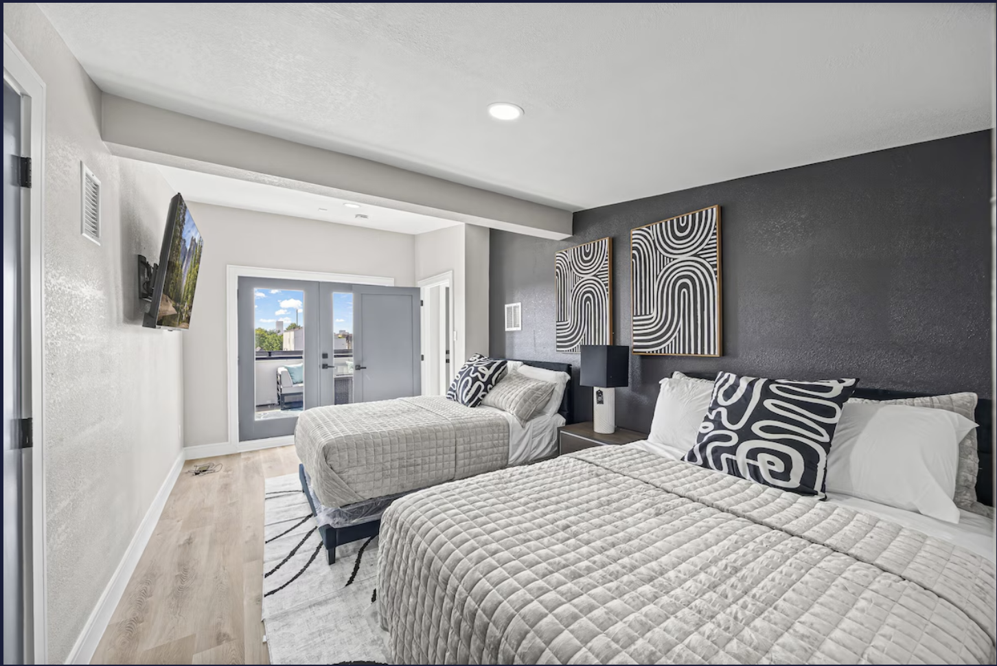 A modern bedroom with two beds featuring beige quilted bedspreads and black-and-white abstract pillows. A black accent wall behind the beds has two framed geometric artworks. There is a nightstand with a black lamp between the beds and a flat-screen 