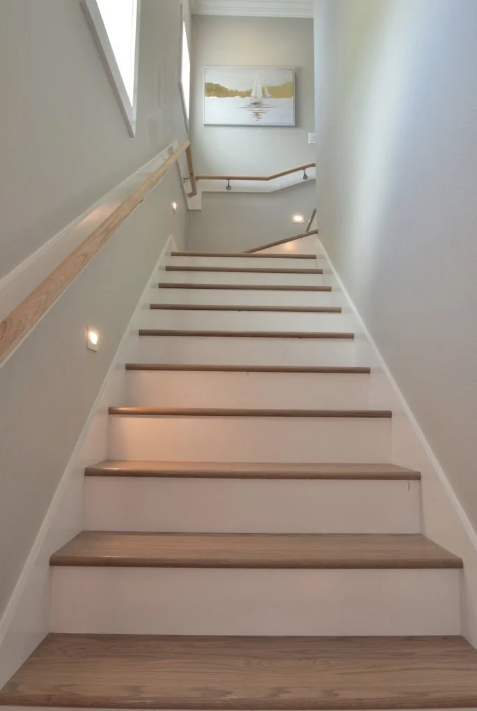 Interior view of a staircase with wooden steps, wall-mounted lights along the side, a wooden handrail on the left, and a framed painting of a sailboat on the upper wall.