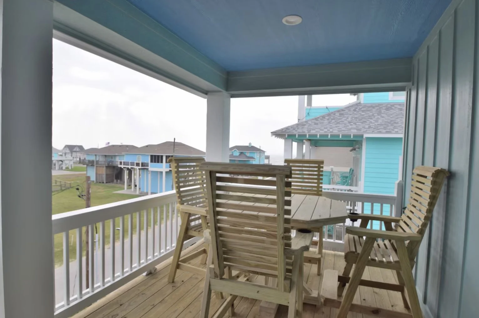 Balcony with wooden table and five wooden chairs, overlooking a neighborhood with colorful beach houses.