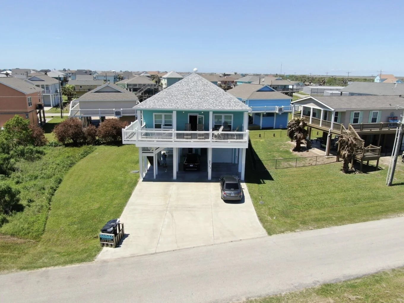 Aerial view of a coastal house with a turquoise exterior, white balcony, garage, and driveway, surrounded by other houses and greenery.
