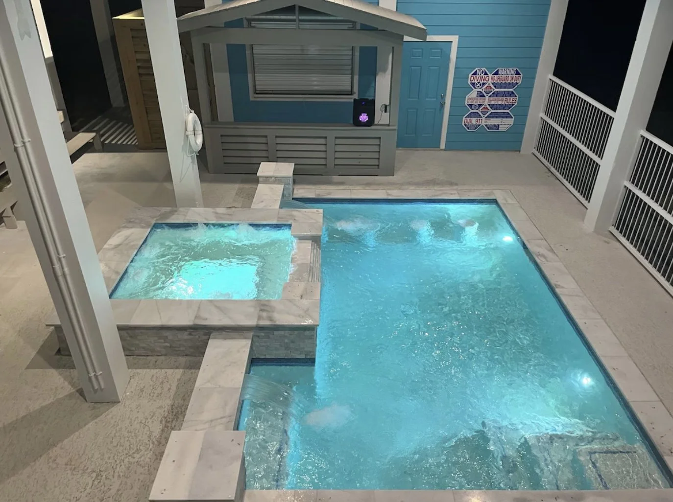 Indoor pool area with a small hot tub connected to a larger swimming pool, illuminated with underwater lights, and a blue wooden wall with signs about diving rules.