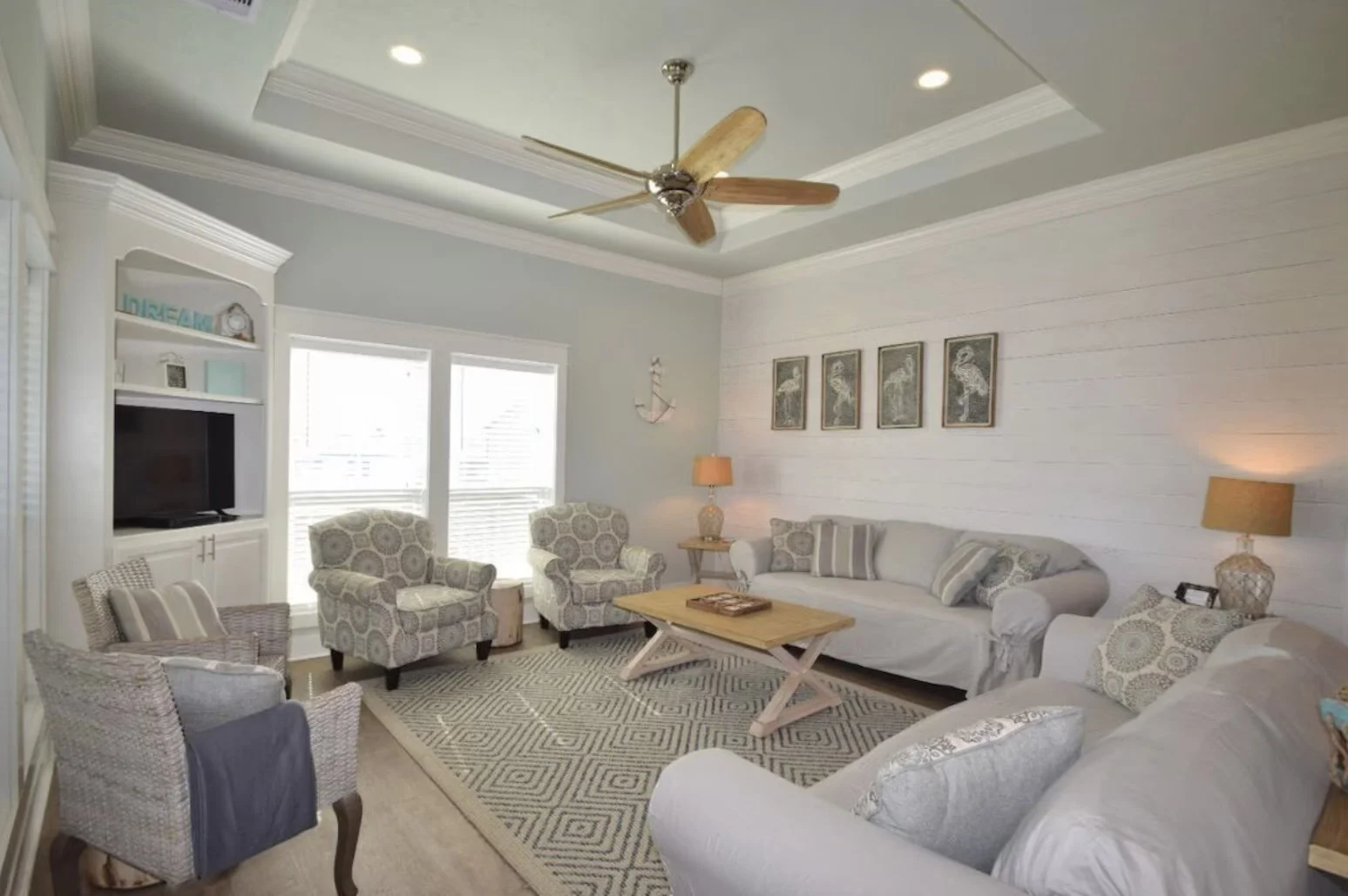Coastal themed living room with white walls, beige upholstered sofas, patterned armchairs, wooden coffee table, framed beach bird artwork, ceiling fan, and large windows with blinds.