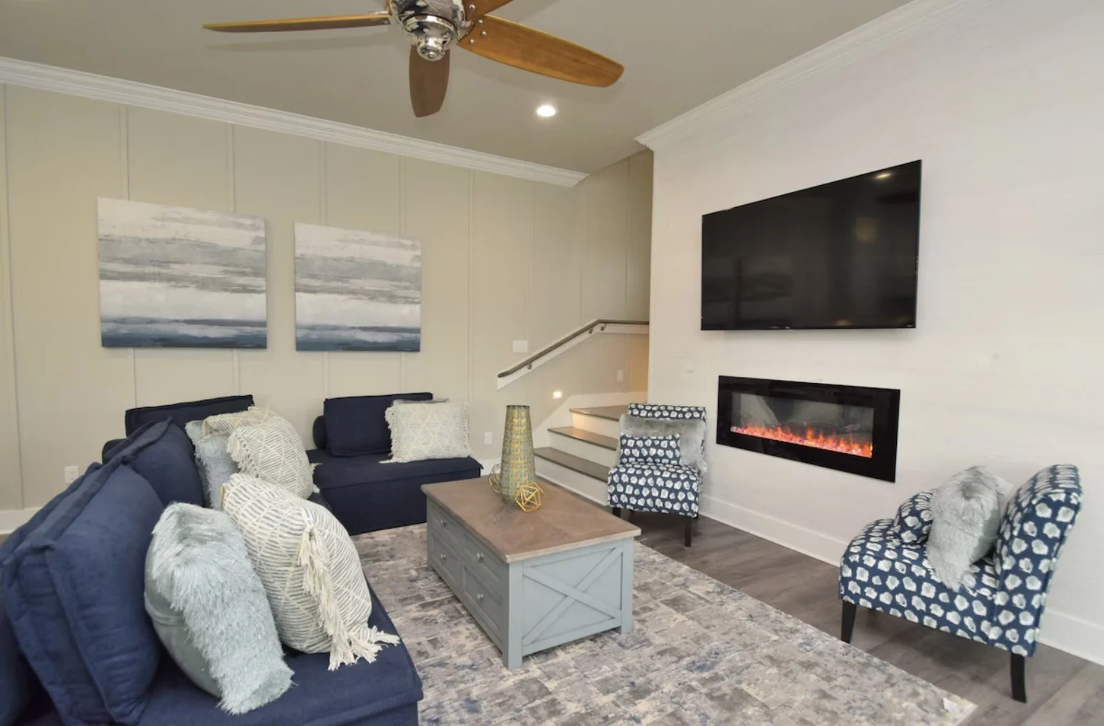 Living room with navy blue sofas and patterned accent chairs arranged around a wooden coffee table. Wall art features two abstract paintings in shades of blue and gray. A modern fireplace with a TV mounted above it is on the right wall. Ceiling fan w