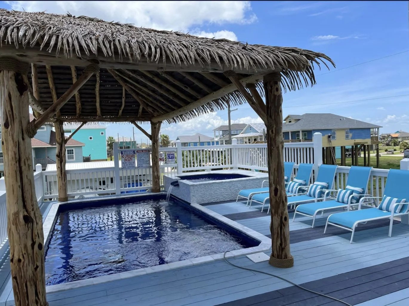 Outdoor pool area with a thatched-roof tiki hut, blue lounge chairs with striped pillows, and a fence with signs, in a coastal neighborhood.