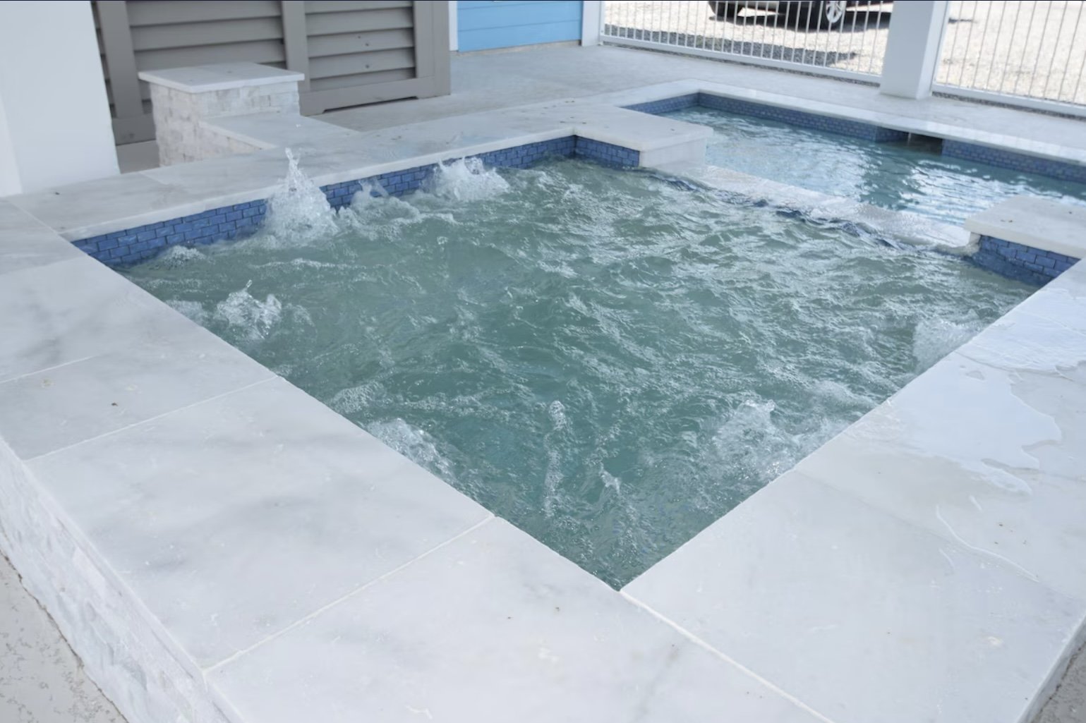 A small outdoor hot tub with bubbling water, surrounded by white stone tiles, with a fence and a blue building in the background.