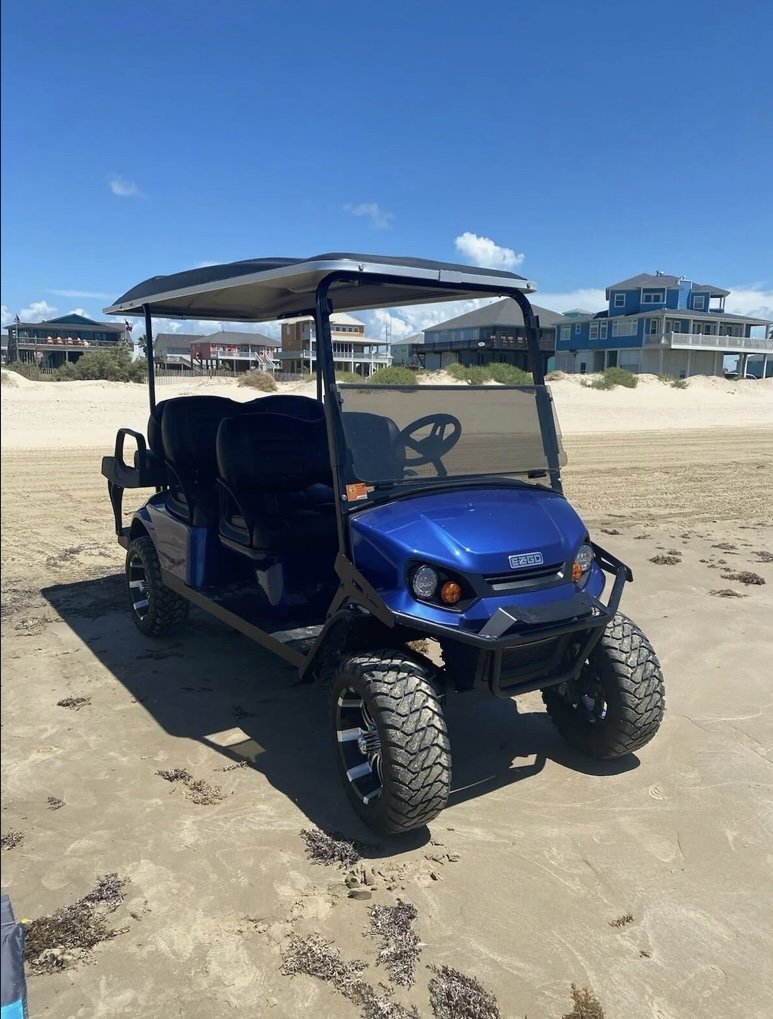 Blue electric golf cart parked on sandy beach with houses in the background under a clear blue sky.