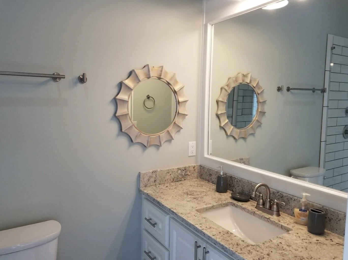 Bathroom with a granite countertop vanity, a large mirror, decorative wall mirrors, a soap dispenser, and a towel rack on a light-colored wall.