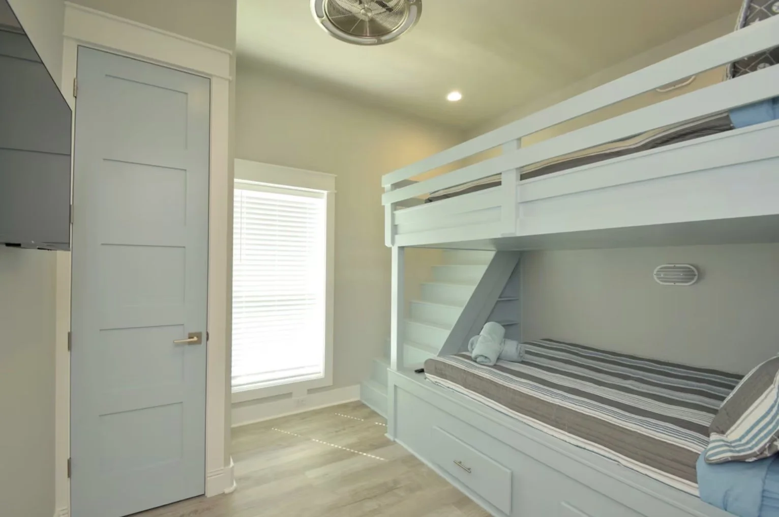 A small room with a built-in bunk bed, a window with blinds, and a closed door painted light blue. The bunk bed has striped bedding and towels, and there is a small drawer underneath the lower bed. The room has light-colored walls, wood flooring, and