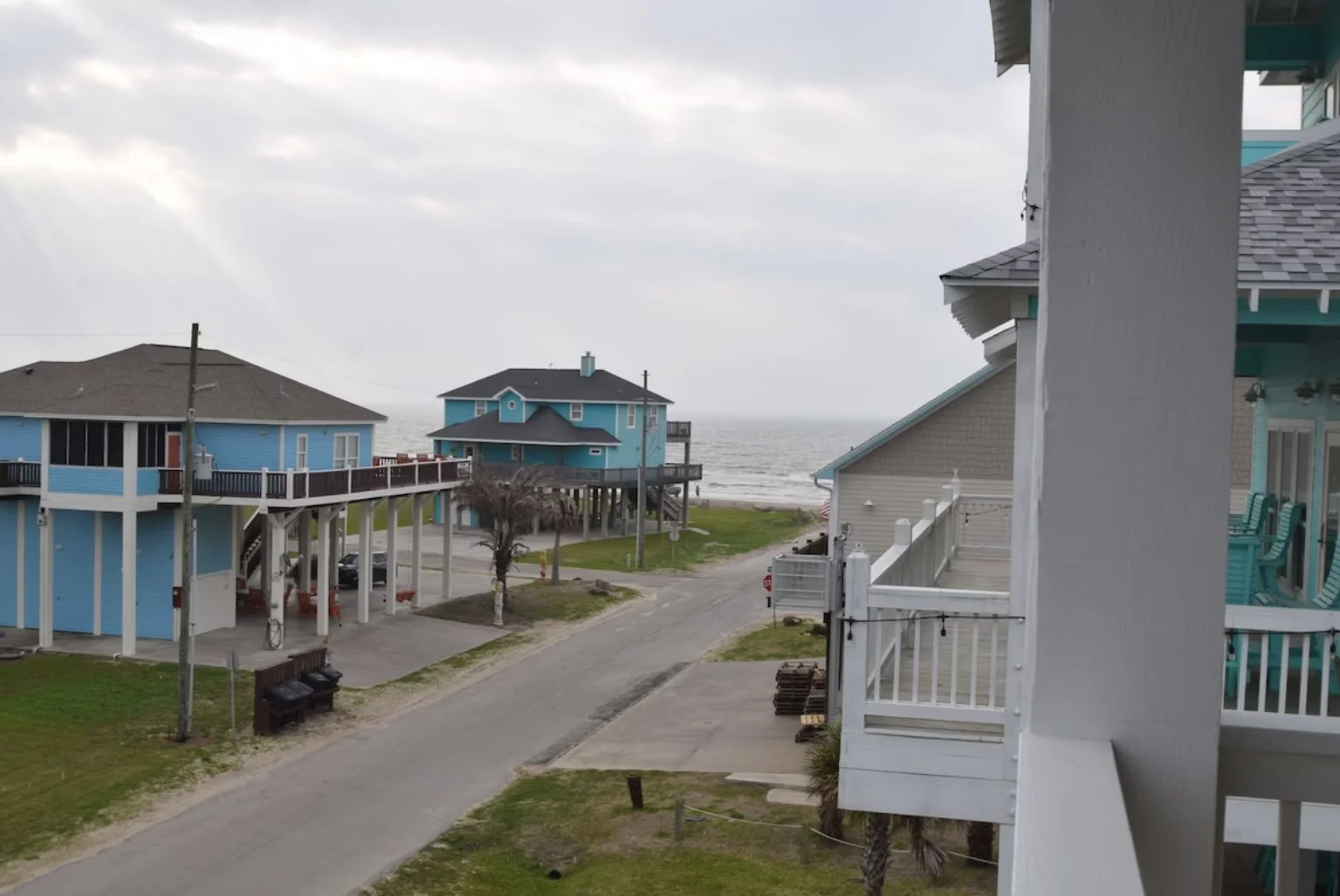 View of beach houses with stilted blue homes overlooking the ocean, a dirt road, and a cloudy sky.