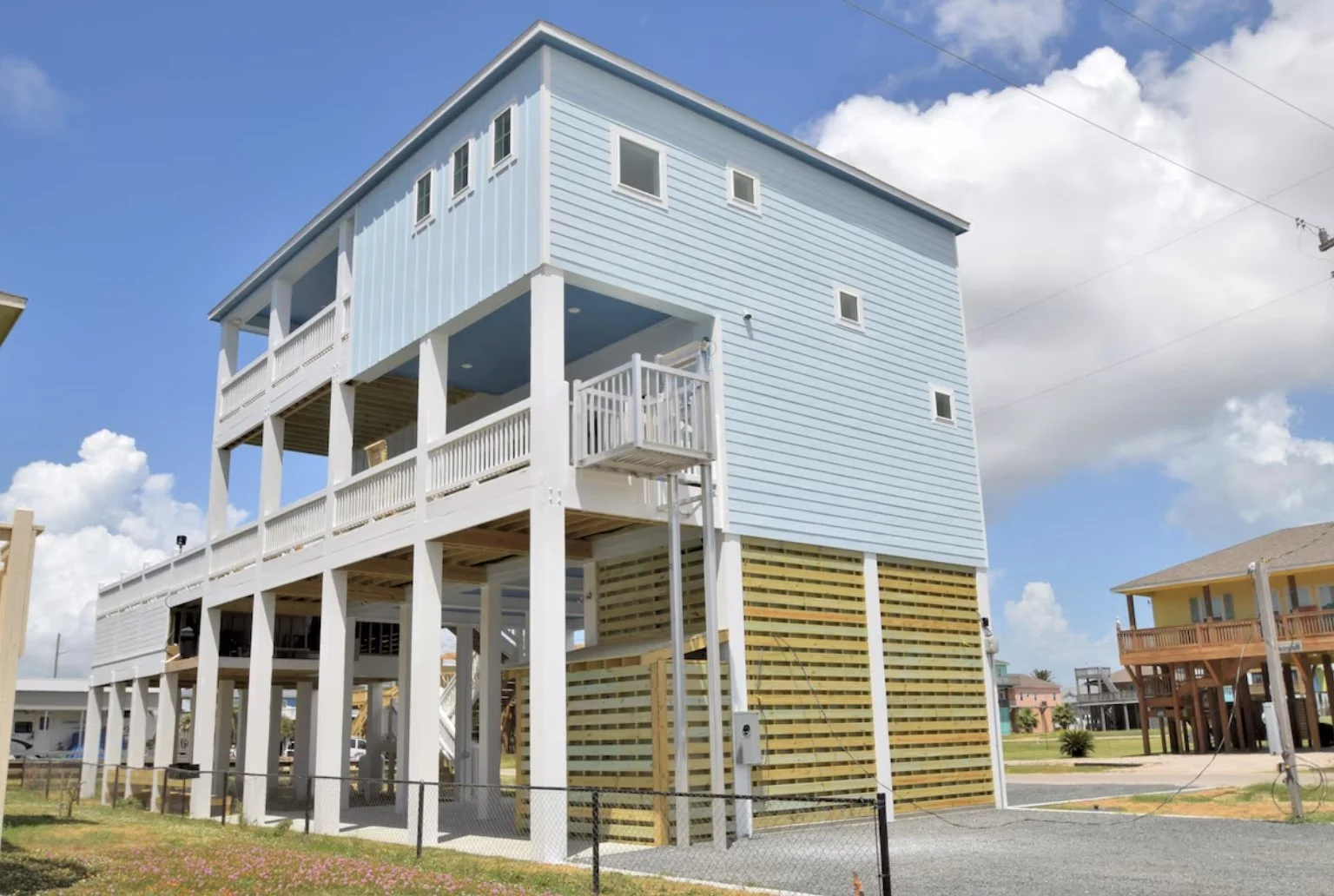 A three-story beach house painted in light blue with white trim, elevated on stilts with an open ground level and a large balcony on the second floor. There are small windows on the upper floors and a staircase visible on the side. The house is situated in a coastal area with other similar houses nearby, under a partly cloudy sky.