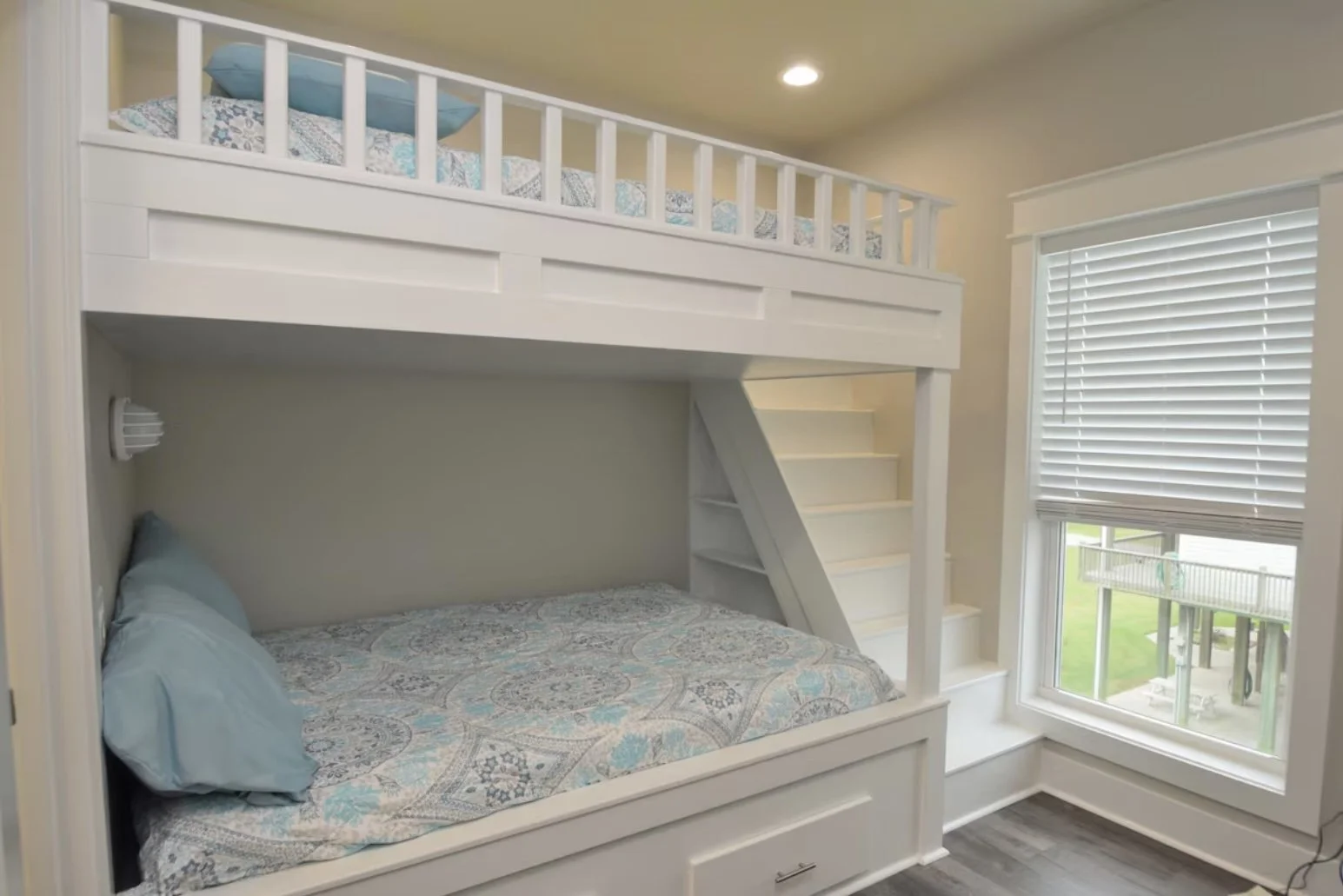 Bunk bed with a lower bed dressed with a blue pillow and patterned bedspread, and an upper bed with a blue pillow and patterned bedding, near a window with blinds, in a bright room with hardwood floors.