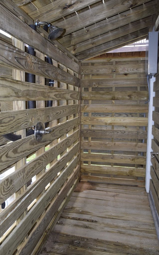 Outdoor wooden shower with a showerhead and a towel hook, enclosed by slatted wooden walls and a sloped roof.