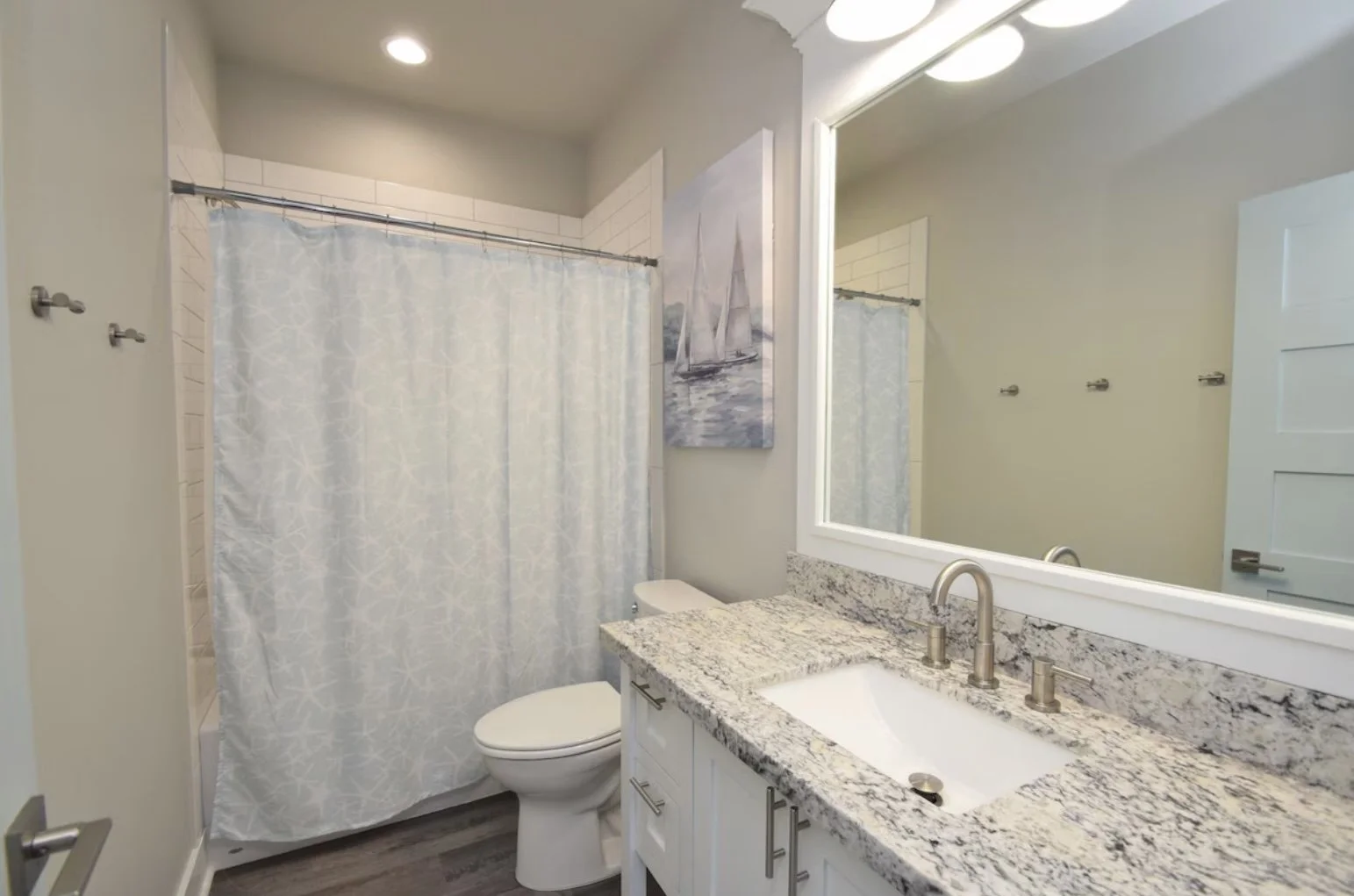 A bathroom with a white vanity with a granite countertop, a large mirror, a sink with a modern faucet, a toilet, and a shower with a curtain. The bathroom has a nautical sailboat painting on the wall.
