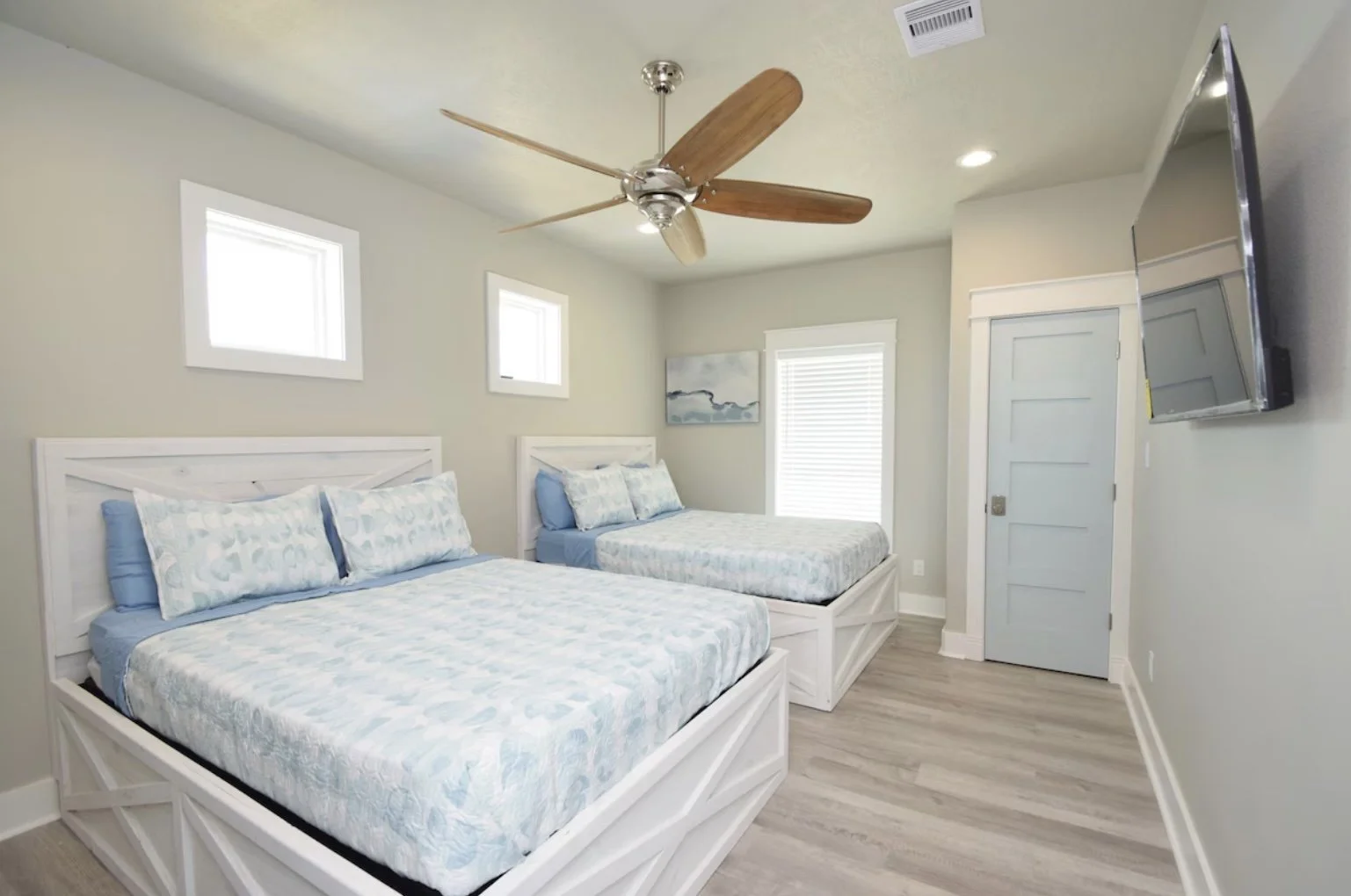 A bedroom with two white beds, light blue and white bedding, a ceiling fan with wooden blades, two small windows, a door, a wall-mounted TV, and a window with blinds.