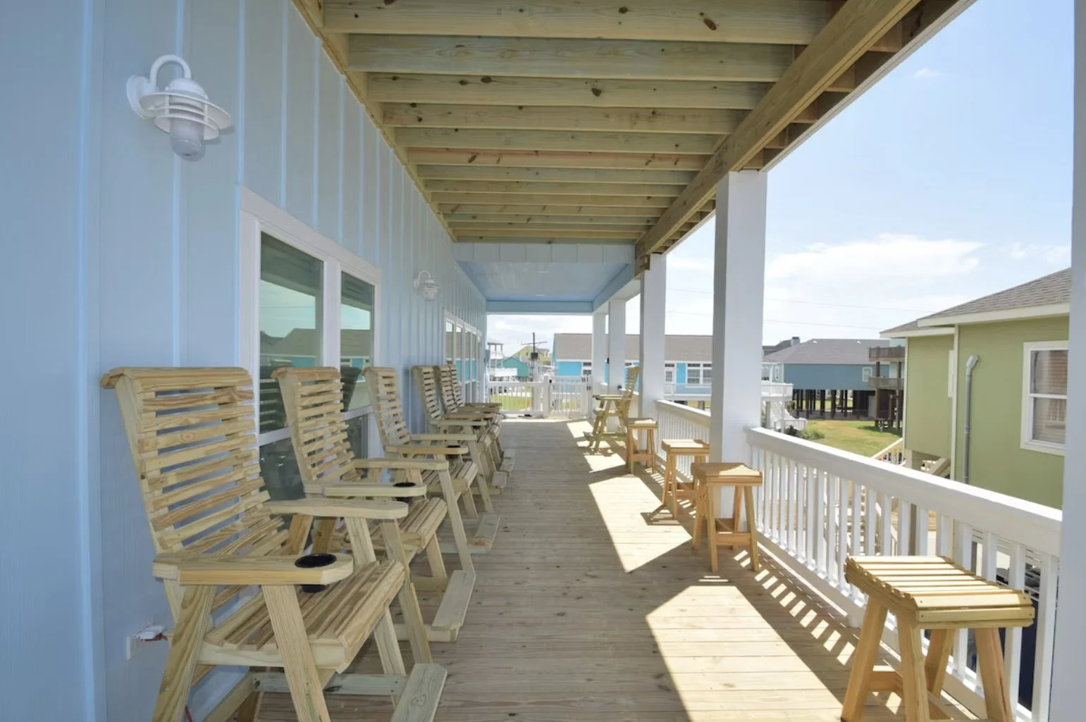 View of a spacious porch with several wooden chairs and small tables, blue siding house, white railing, and neighboring houses in the background on a sunny day.