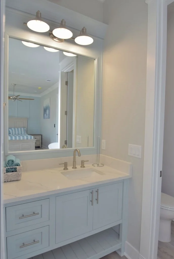 Bathroom vanity with a large mirror, three light fixtures above, a white countertop with a sink, and a basket with rolled towels. Part of a bedroom with a bed and a painting is visible through the mirror reflection.