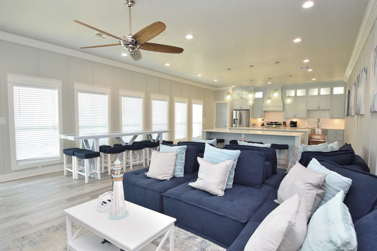 Open concept living room with navy blue sectional sofa, multiple light-colored pillows, white coffee table with lighthouse decor, kitchen with gray cabinets, white backsplash, stainless steel refrigerator, pendant lights, and a bar counter with black