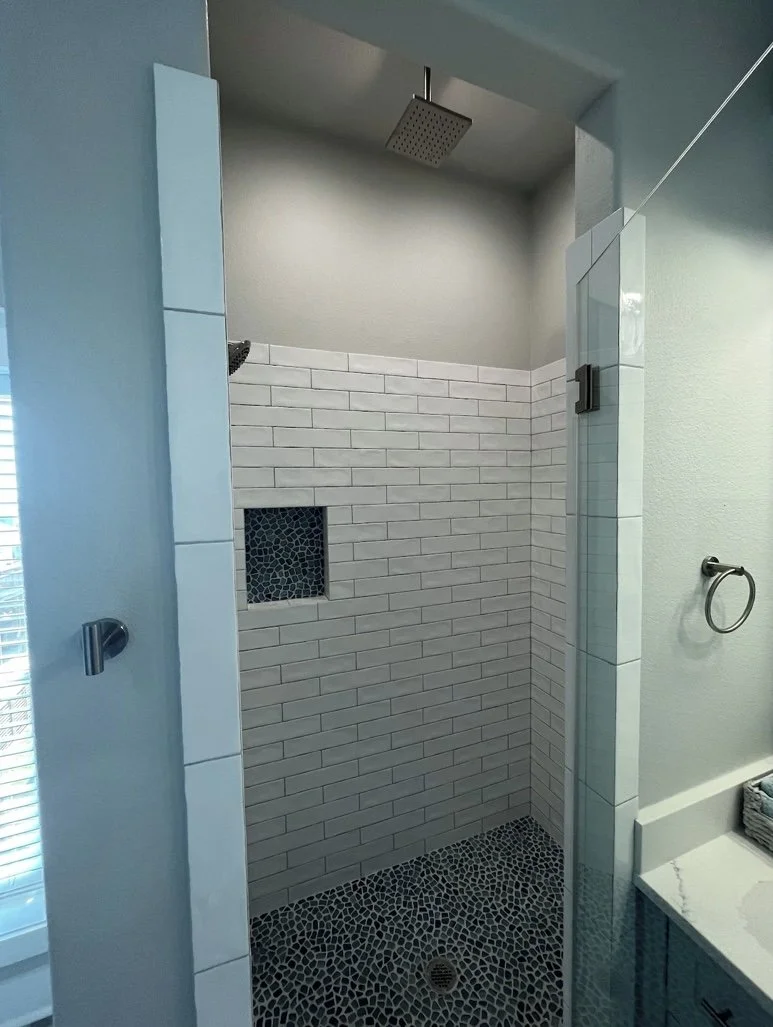 Modern walk-in shower with white subway tiles, pebbled black floor, square overhead shower head, small mosaic accent wall, and glass door.