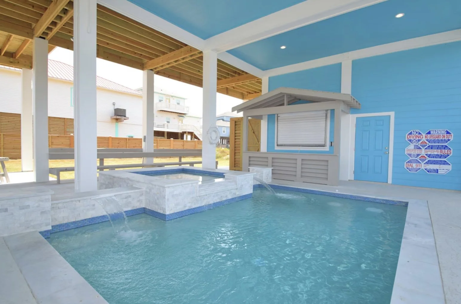 Indoor swimming pool area with a small spa, blue walls, and wooden accents, featuring a window with a roller shutter, a door, and a wall with safety signs.