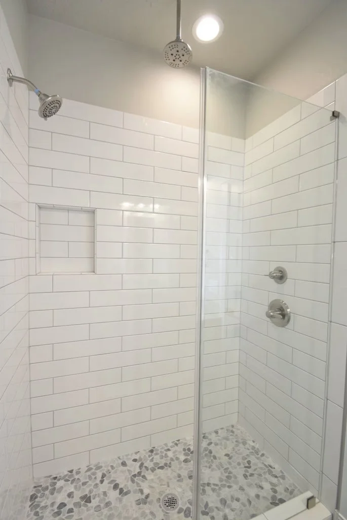 Modern walk-in shower with white subway tiles, pebble stone floor, a built-in niche, and dual showerheads, in a bathroom with a ceiling light.