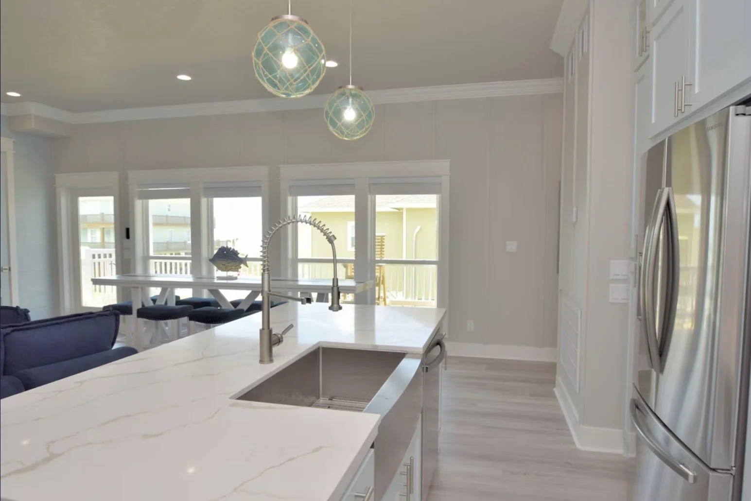 Modern kitchen with white marble island, stainless steel refrigerator, light wood flooring, and large windows with a balcony view. Hanging pendant lights above the island and a dual-function faucet at the sink.