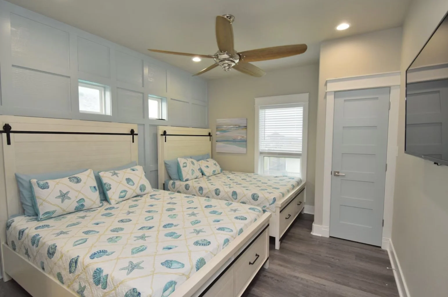 Beach-themed bedroom with two twin beds featuring seashell and starfish bedding, a ceiling fan, a window with blinds, and a wall-mounted TV.