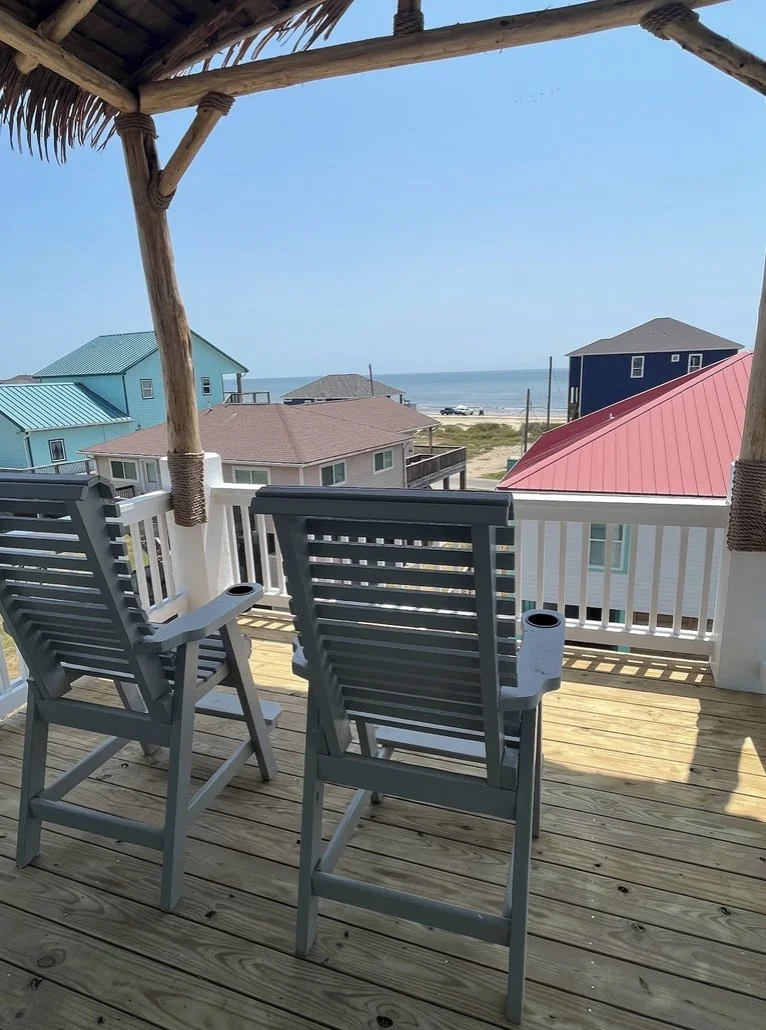 Beach view from a wooden deck with two chairs, overlooking colorful houses and the ocean.