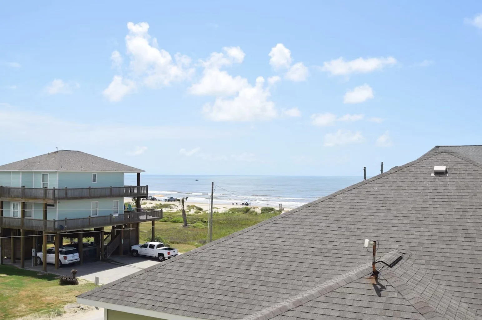 View of a beach with ocean, sandy shore, and clouds, seen from rooftops of beachfront buildings with parked cars, grassy area, and palm trees.