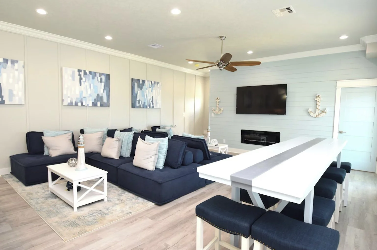 Bright living room with navy sectional sofa, white pillows, abstract blue and white artwork, light wood flooring, and nautical decor including two anchor wall hangings, a wall-mounted flat-screen TV, a white fireplace, and a white bar-height table wi
