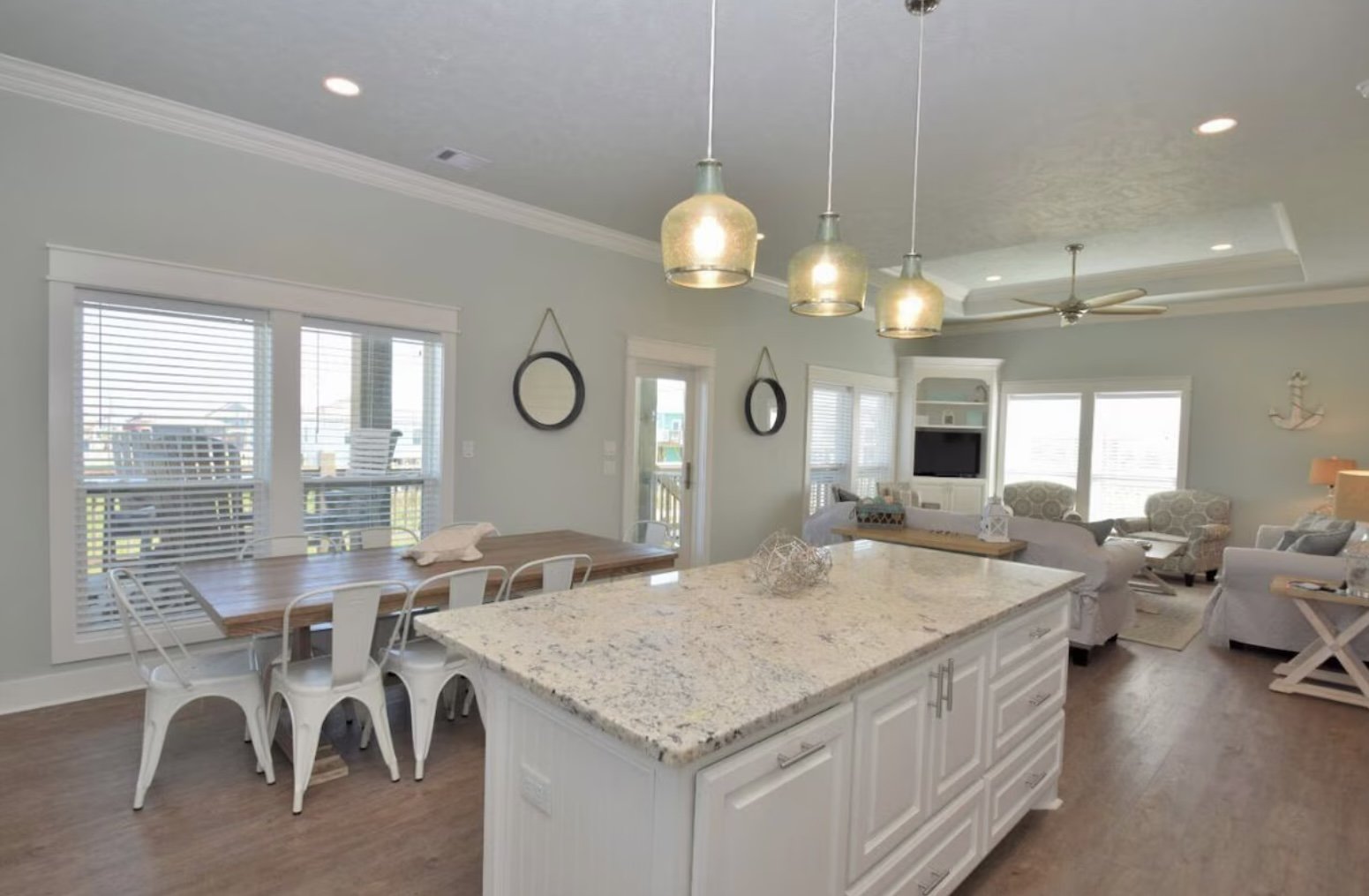 Open-concept living and kitchen area with a large granite island, white cabinets, pendant lighting, a dining table with white chairs, multiple windows with blinds, and a seating area with sofas and chairs.