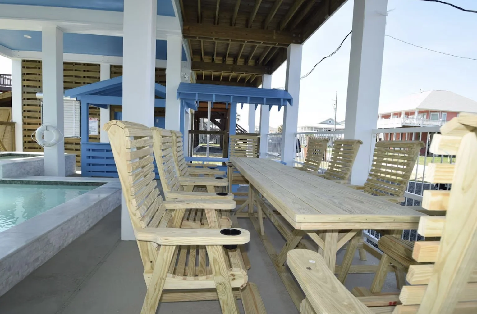 Outdoor covered patio with a long wooden table, wooden chairs, and a small hot tub or pool in the foreground. There is a blue gazebo and a playhouse in the background.