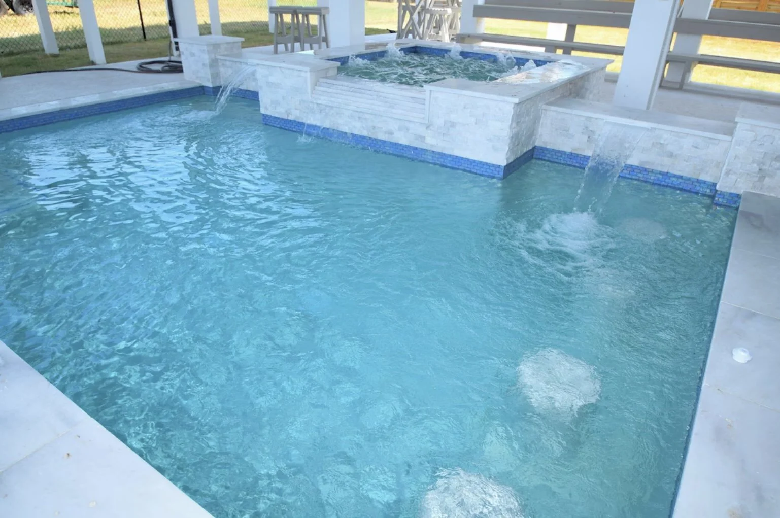 A backyard swimming pool with a hot tub attached, featuring bubble jets and a stone border. The pool area is surrounded by a wooden deck and a white fence.