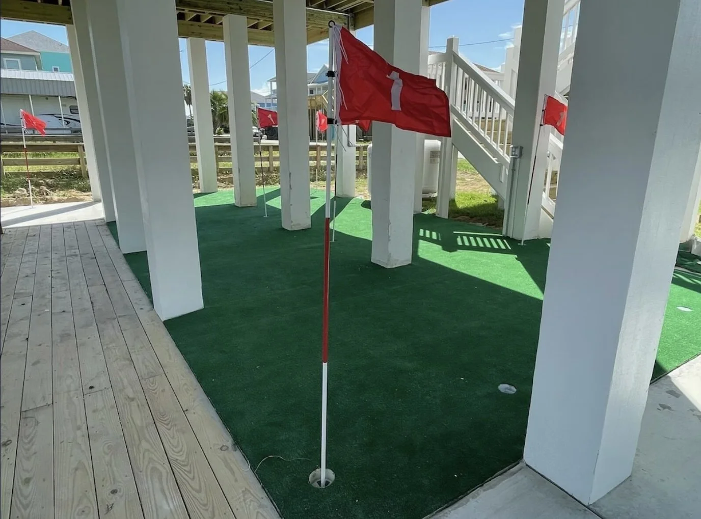 A mini golf hole with a red flag marked with the number one, white posts, green artificial turf, and a wooden deck surface nearby, under a shaded structure.