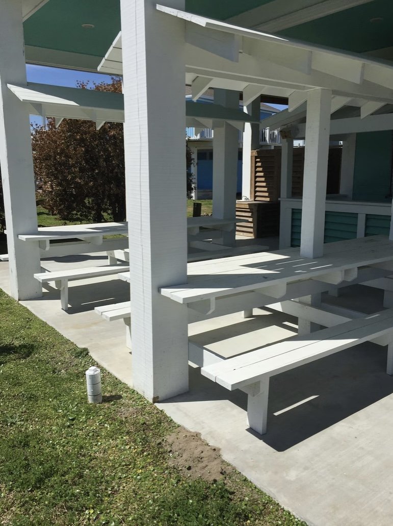 White wooden picnic tables and benches under a building overhang, with a grassy area and a tree in the background.