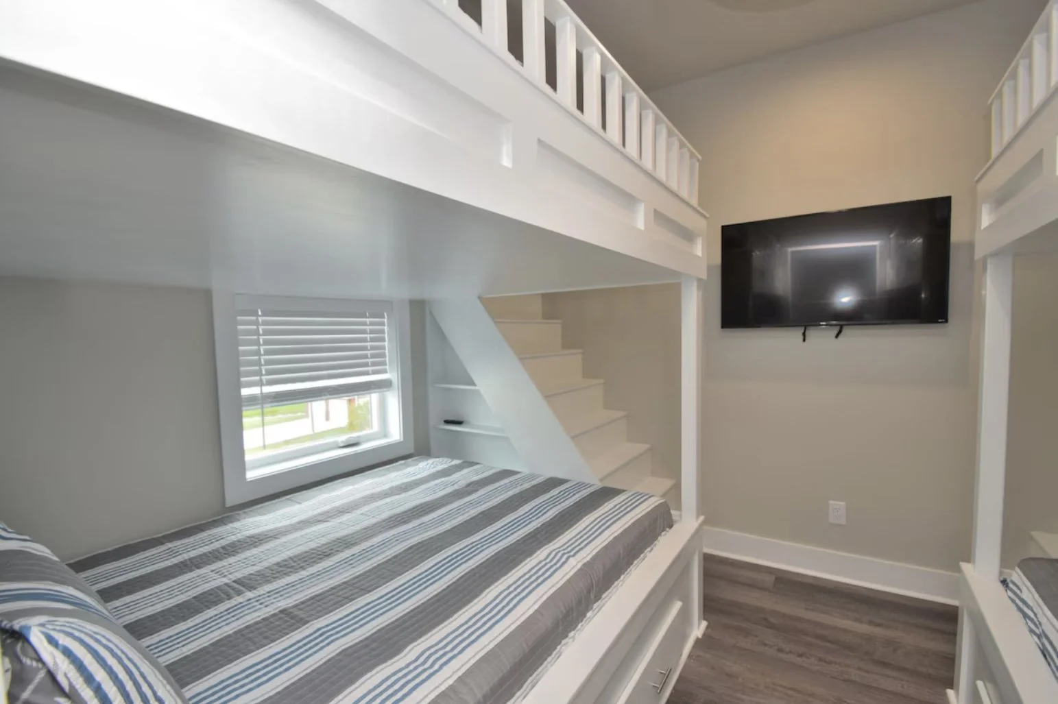 A small bedroom with a built-in bunk bed featuring striped bedding, a window with blinds, a staircase, a wall-mounted TV, and neutral-colored walls and flooring.