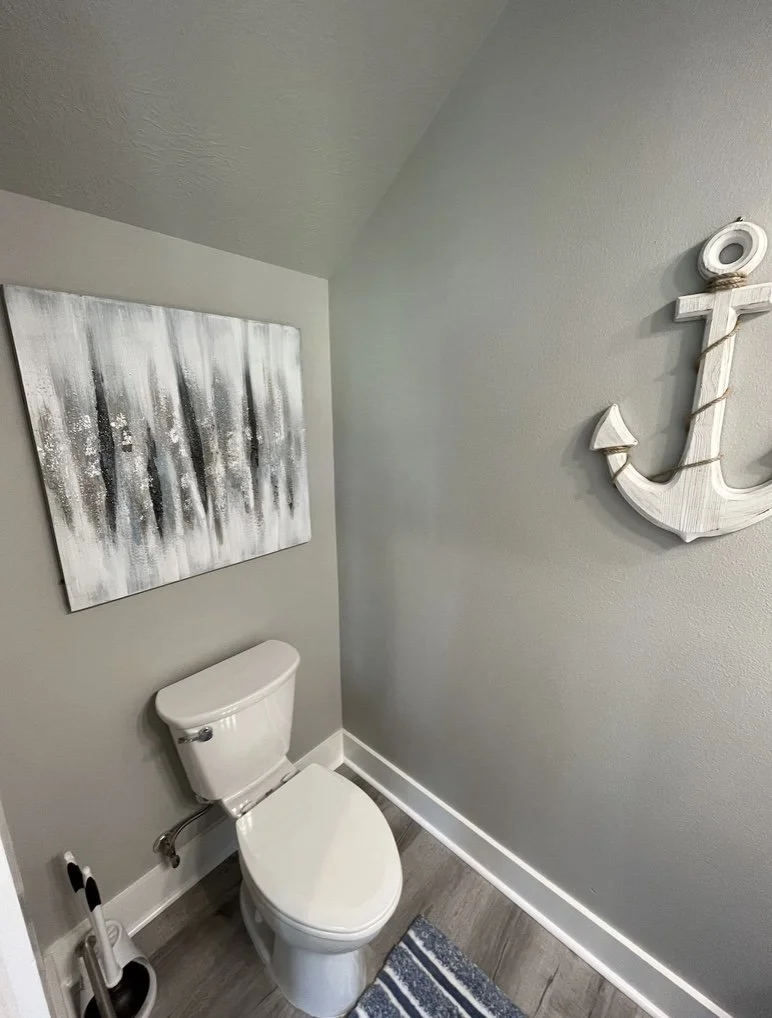 Small bathroom with a white toilet, abstract ocean-themed artwork on the wall, a nautical anchor decoration, and a striped rug on wood-look flooring.
