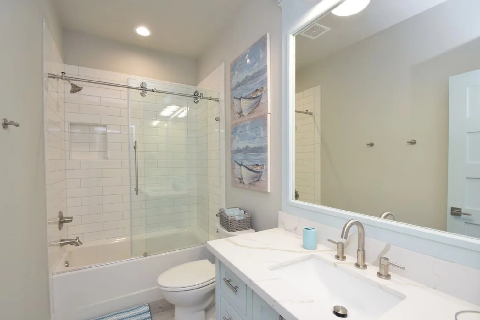 Bathroom with a shower, bathtub, toilet, large mirror, and white vanity with a sink, blue soap dispenser, and wall art of boats and water.
