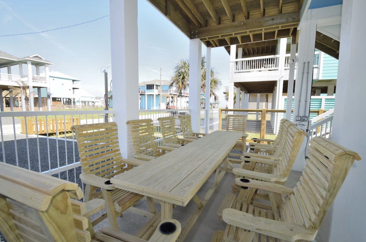 Outdoor patio area with wooden table and chairs, surrounded by white railings, with beach houses and palm trees in the background.