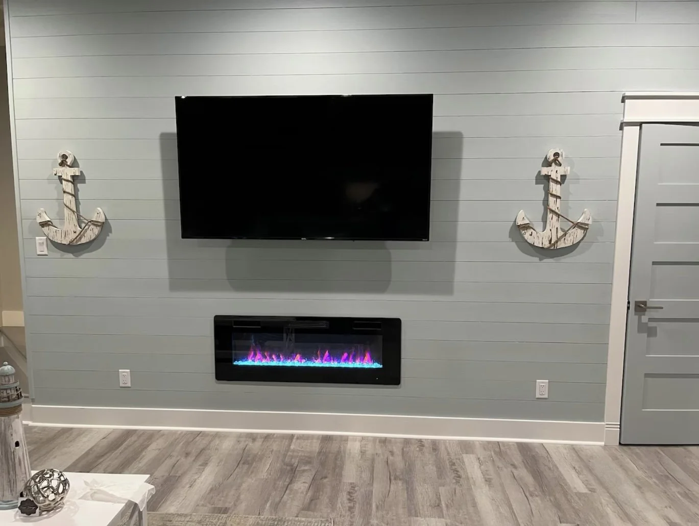 Living room with a wall-mounted flat-screen TV, two nautical anchor decorations, and an electric fireplace with purple flames, light gray wall paneling, and light wood flooring.