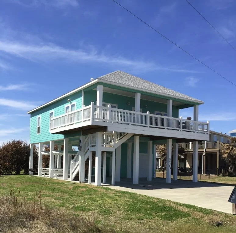 A two-story beach house with turquoise siding, white trim, and a gray shingled roof. The house is raised on stilts with a staircase leading to a large front porch with white railings.