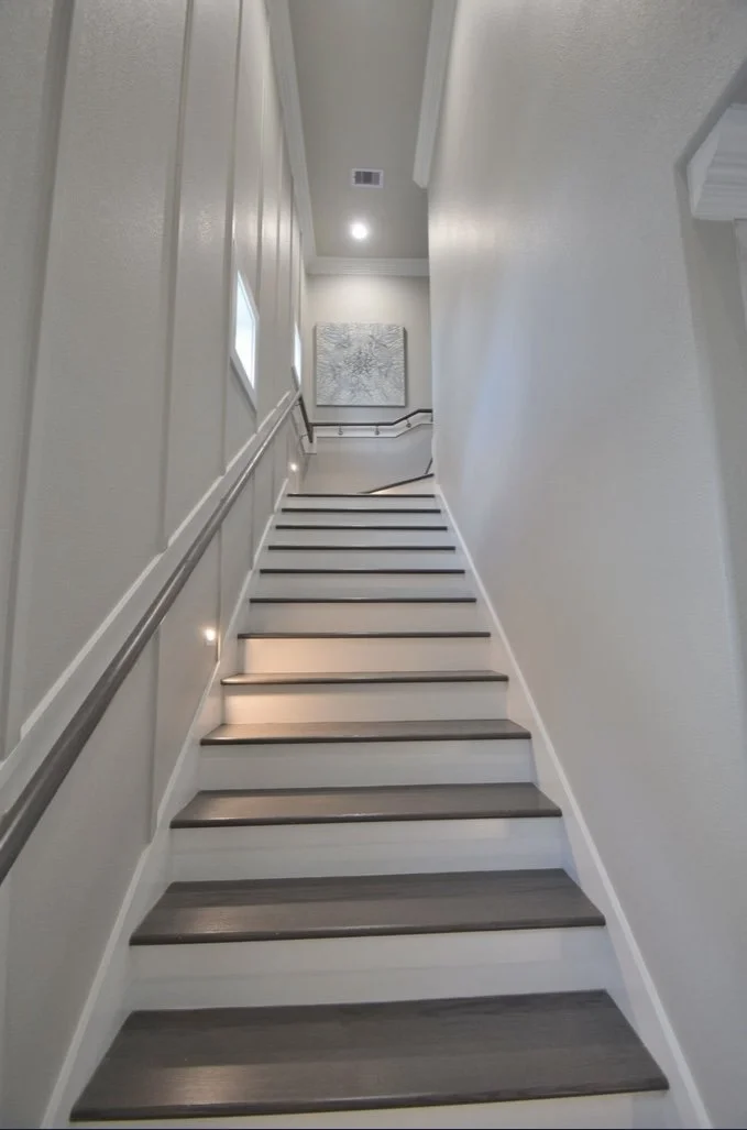 A downward view of a modern staircase with dark wood steps, white risers, and a black handrail on the left side, leading to a brightly lit upper level with artwork on the wall.