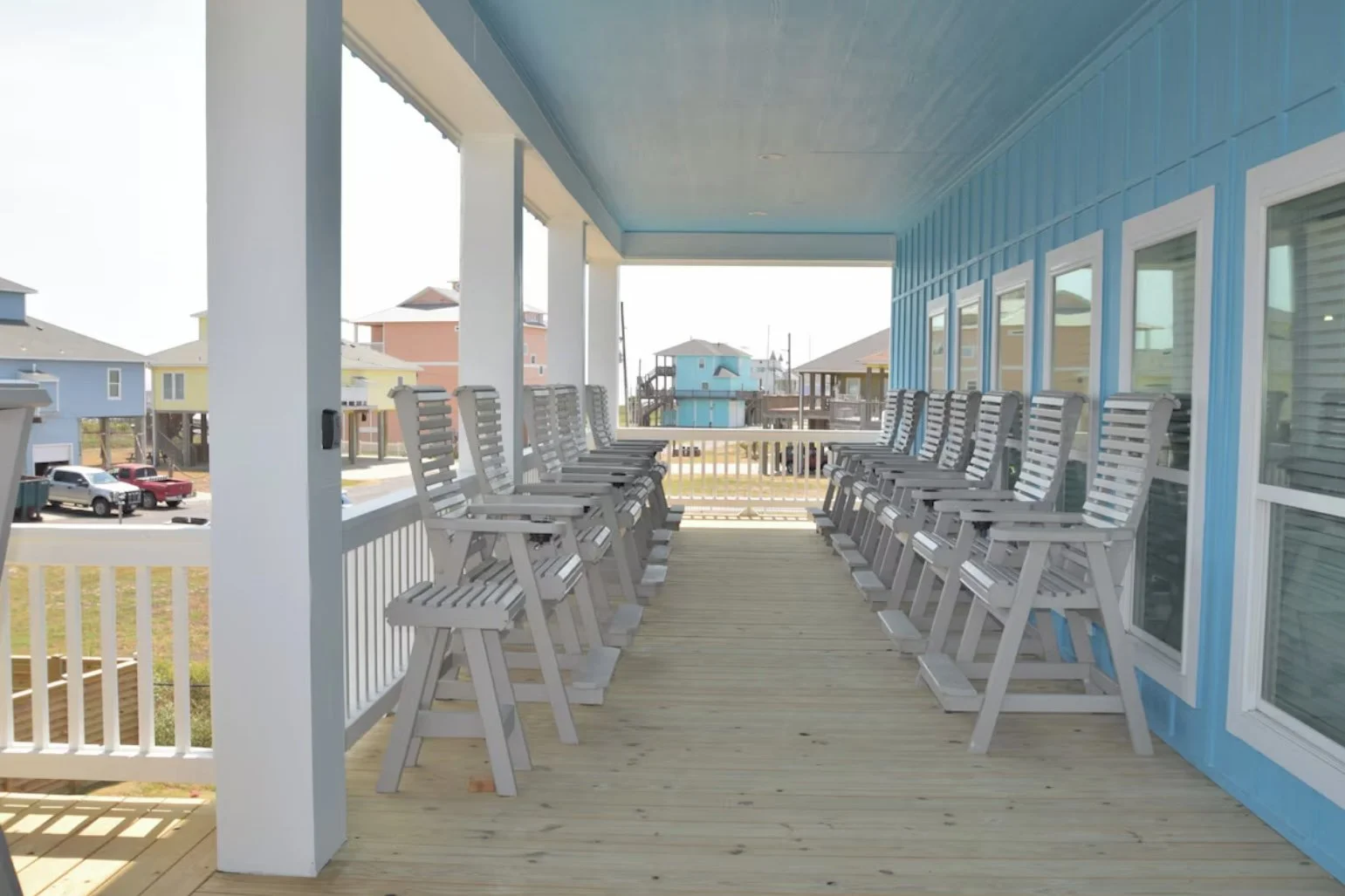 Outdoor balcony with two rows of white wooden chairs facing each other, overlooking colorful houses and a parked car.