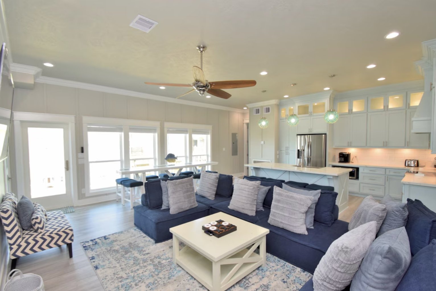 Open-concept living room and kitchen with navy sectional sofa, armchair with chevron pattern, coffee table, bar-height dining table near windows, white cabinetry, stainless steel refrigerator, and pendant lighting.
