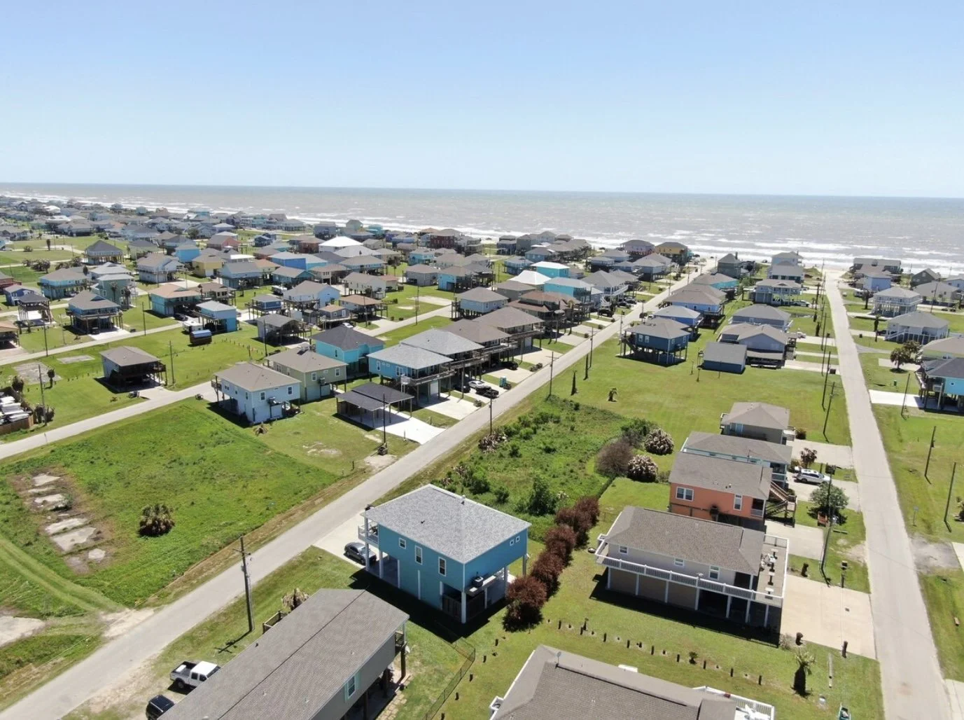 Aerial view of a beachside residential neighborhood with colorful houses, green lawns, paved roads, and the ocean in the background.