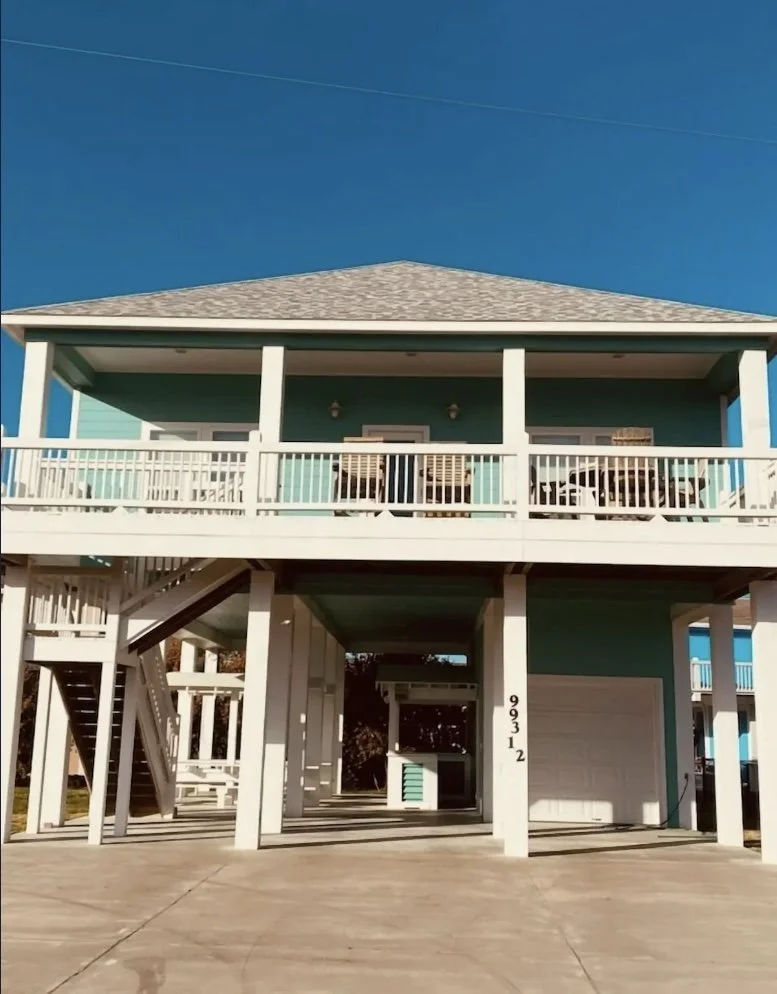 Two-story beach house elevated on stilts with an open parking area underneath, white railings, turquoise exterior, stairs leading to the upper porch, and a clear blue sky overhead.