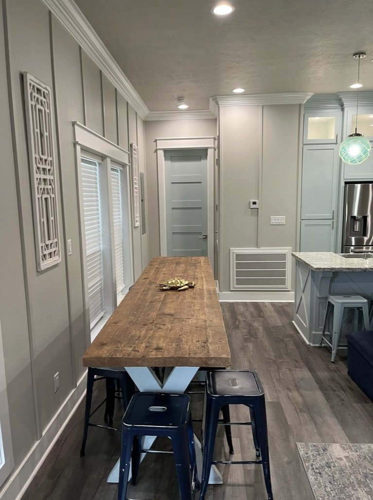 A modern kitchen and dining area with a rustic wooden table, four blue metal stools, white wainscoting, gray paneled walls, and a kitchen counter with a granite top.