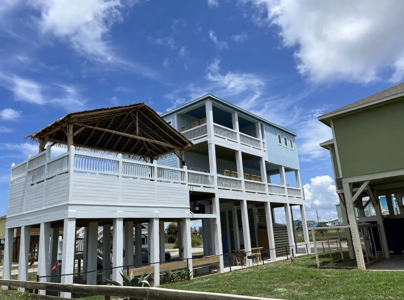 Beach house with multiple decks and a thatched roof gazebo, under a partly cloudy sky.
